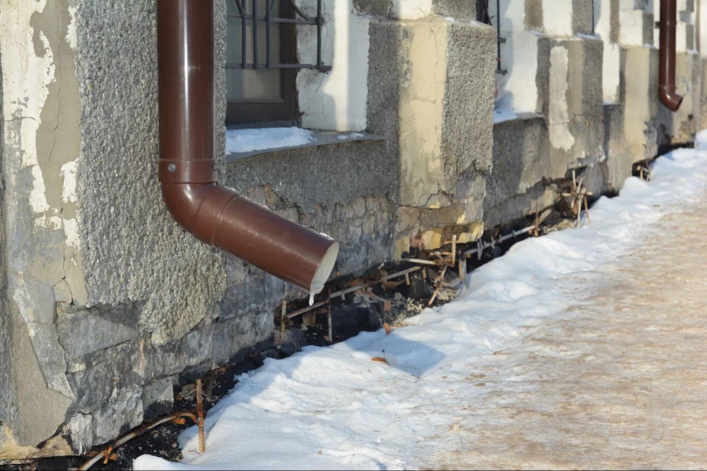 Downspout beside cracked exterior walls and a snow-covered walkway.