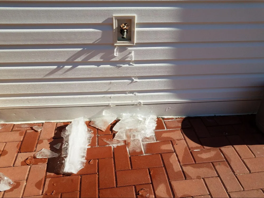 Ice on exterior siding and brick paving beneath an outdoor faucet.